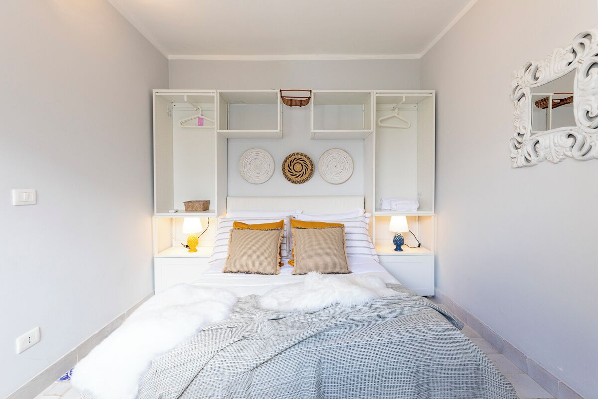 A bedroom with a bed, a white wardrobe, a mirror, and a bedside table with a lamp.
