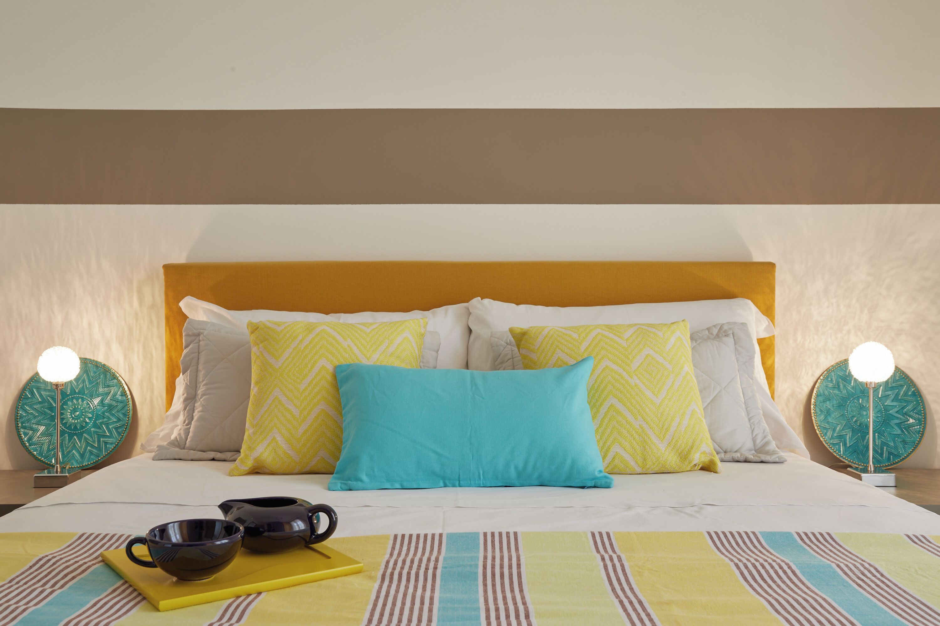 Una cama bien hecha con una colcha a rayas, dos lámparas de noche y una bandeja con tetera y tazas.