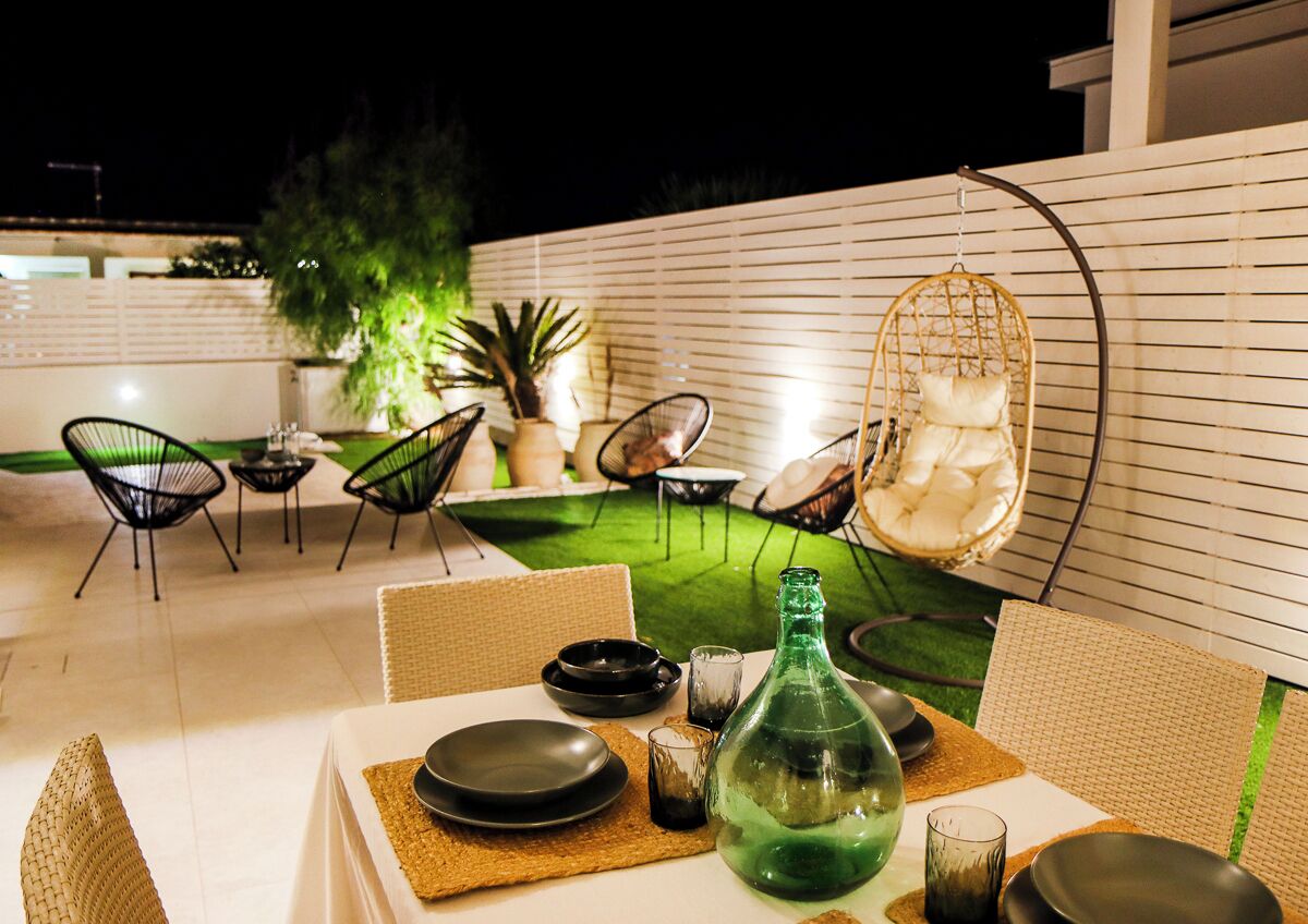 An outdoor dining area with wicker chairs, a hanging chair, and a table set with plates, glasses, and a green bottle.