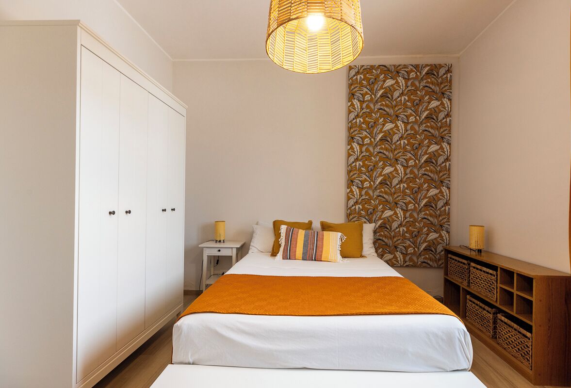 A bedroom with a bed, white wardrobe, bedside table, lamp, and a wall with a leaf pattern.