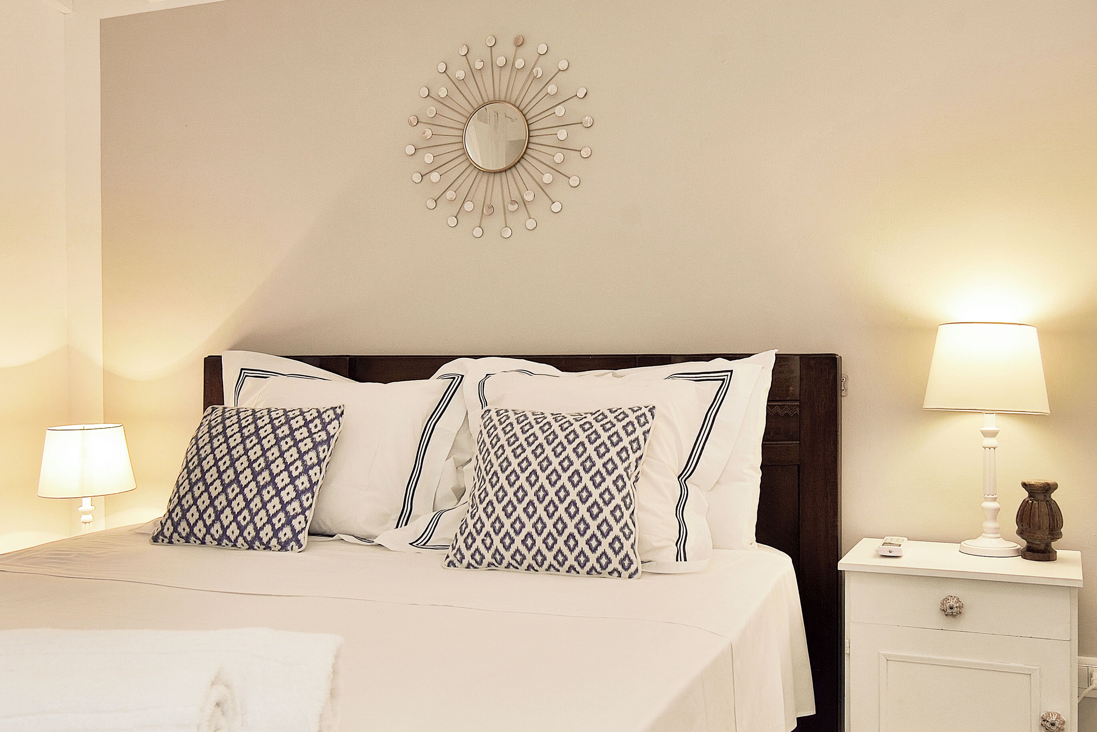 Una cama bien hecha con almohadones estampados, un espejo montado en la pared, lámparas de noche y una mesita de noche con una lámpara y un florero.