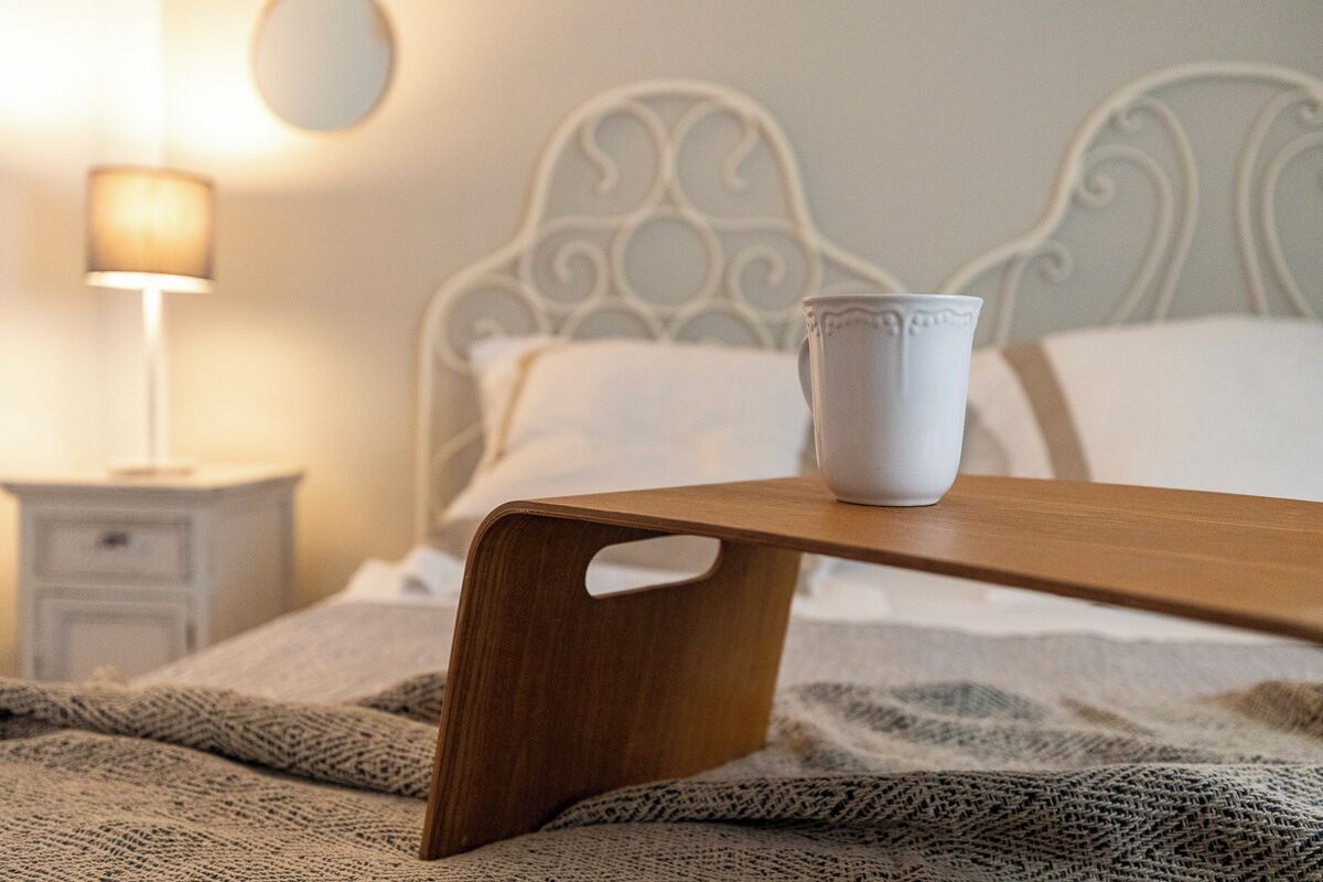 A wooden bedside table with a white mug on it, a white metal bed frame, a white nightstand with a lamp, and a knitted blanket on the bed.