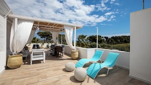 Eine sonnendurchflutete Terrassenanlage im Freien mit Holzdeck, weißen Vorhängen, einem Tisch mit Stühlen und blauen Liegestühlen.