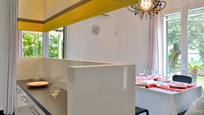 Un comedor moderno con una mesa blanca dispuesta para cuatro personas, una lámpara decorativa y amplias ventanas que ofrecen vista a la vegetación exterior.
