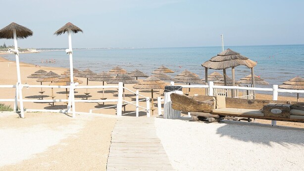 Una playa con un paseo de madera que lleva a un área de descanso con sombrillas de paja.