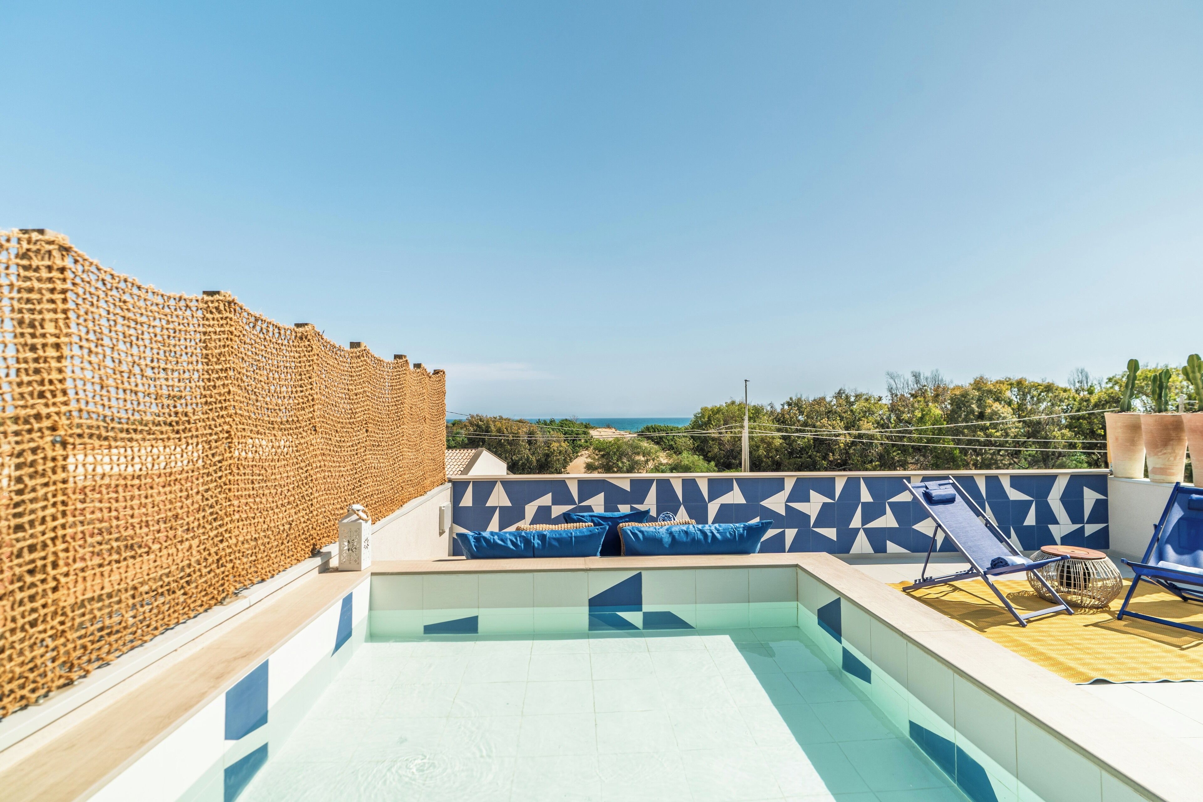 A rooftop pool with blue and white tiles, a wooden deck, and a view of the sea.