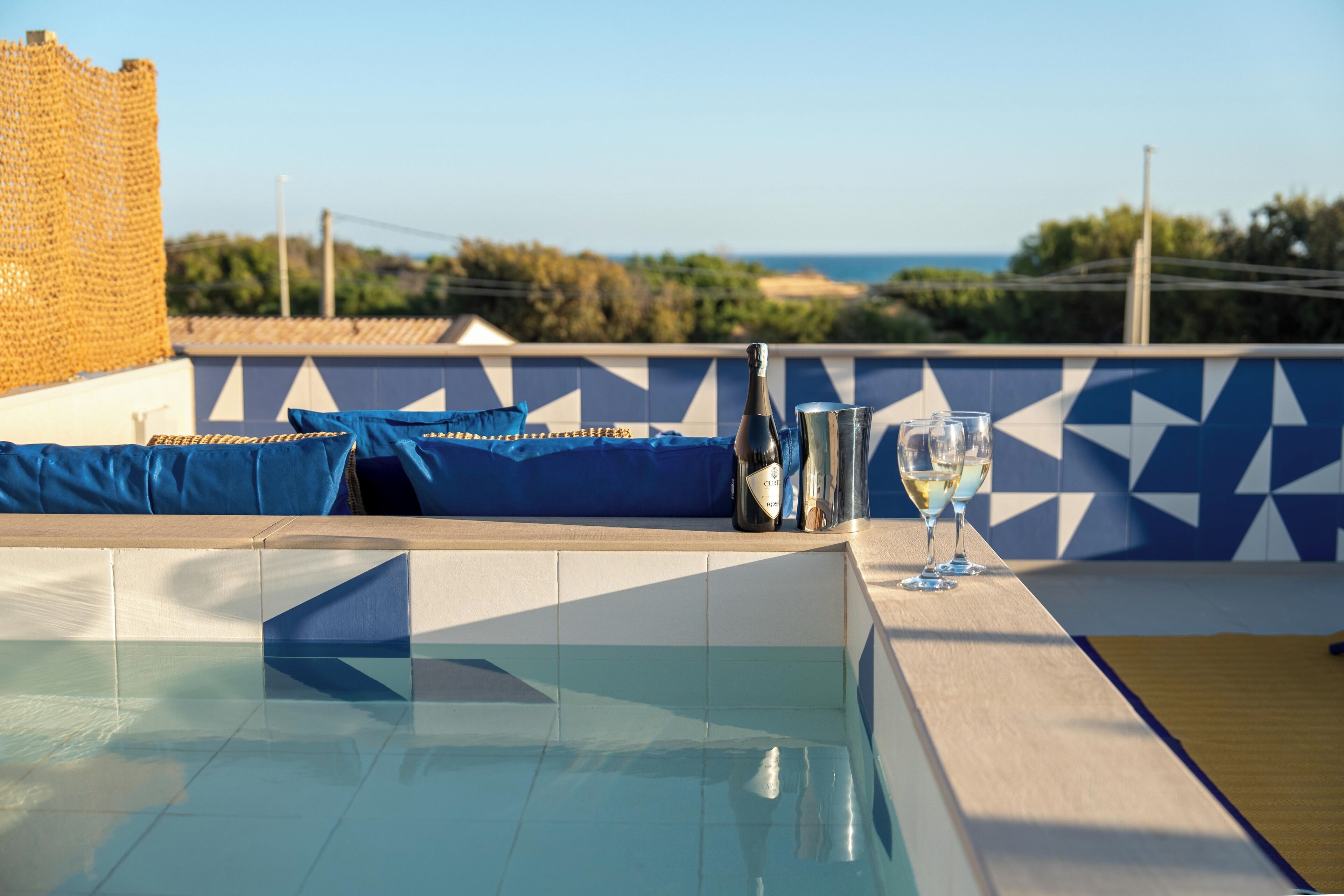 Un espace piscine avec vue sur la mer, un mur recouvert de carreaux formant un motif géométrique, et une table avec des verres et des bouteilles.