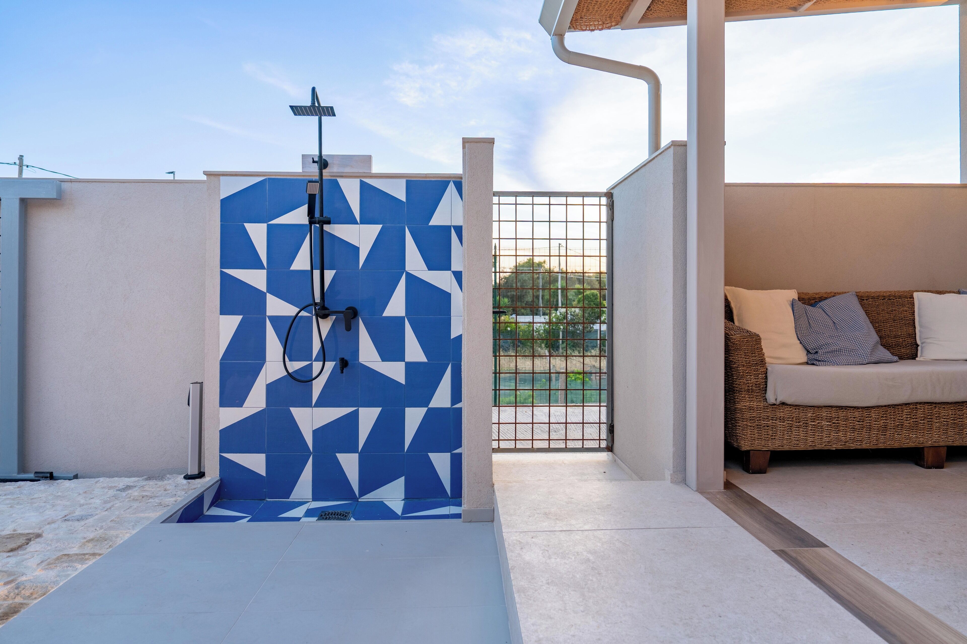 Uma área de chuveiro externa moderna, com padrões geométricos em azulejos, um sofá de vime com almofadas e vista para vegetação através de um portão.