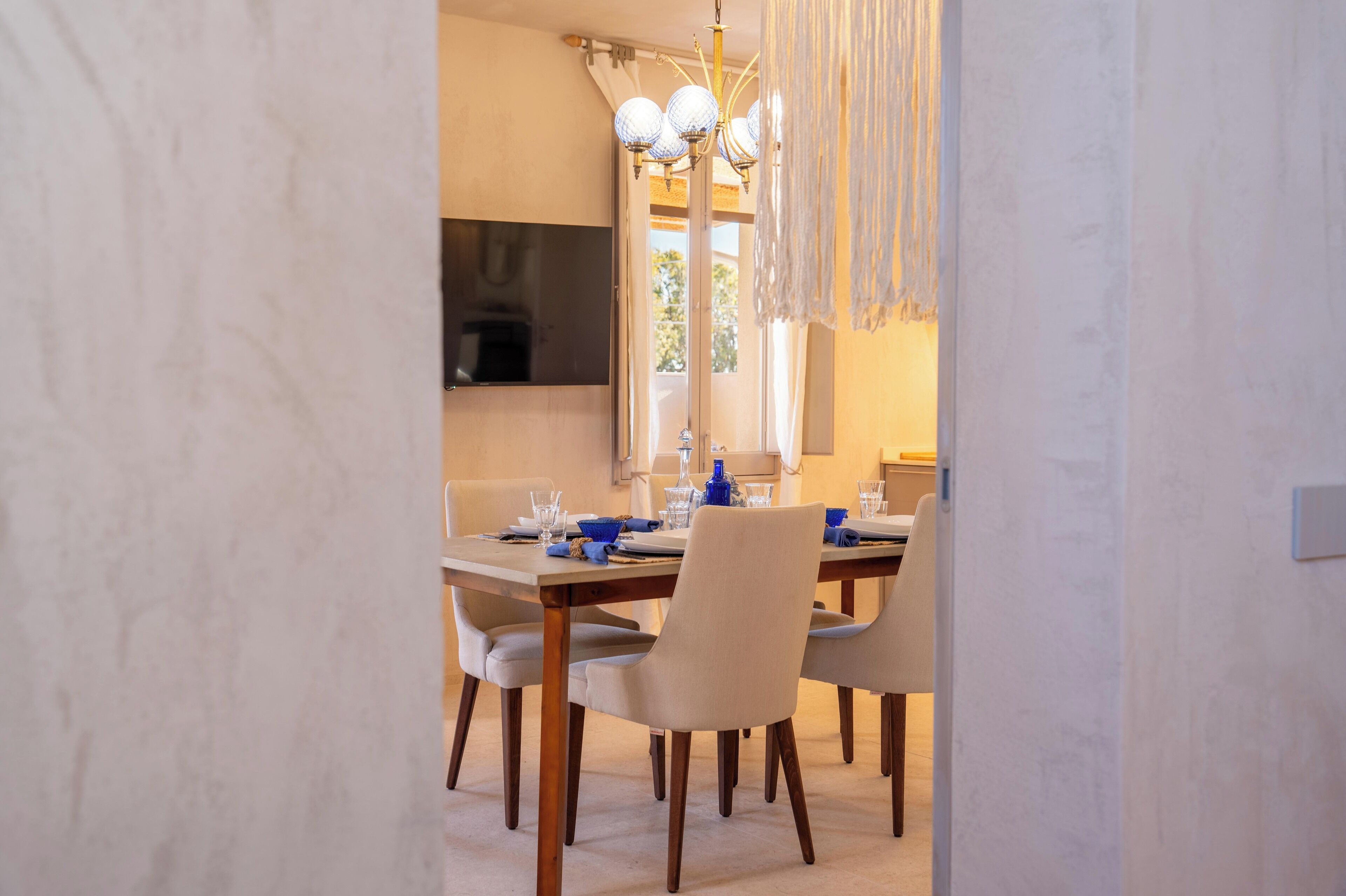 A dining area with a table set for four, a chandelier, and a flat-screen TV mounted on the wall.