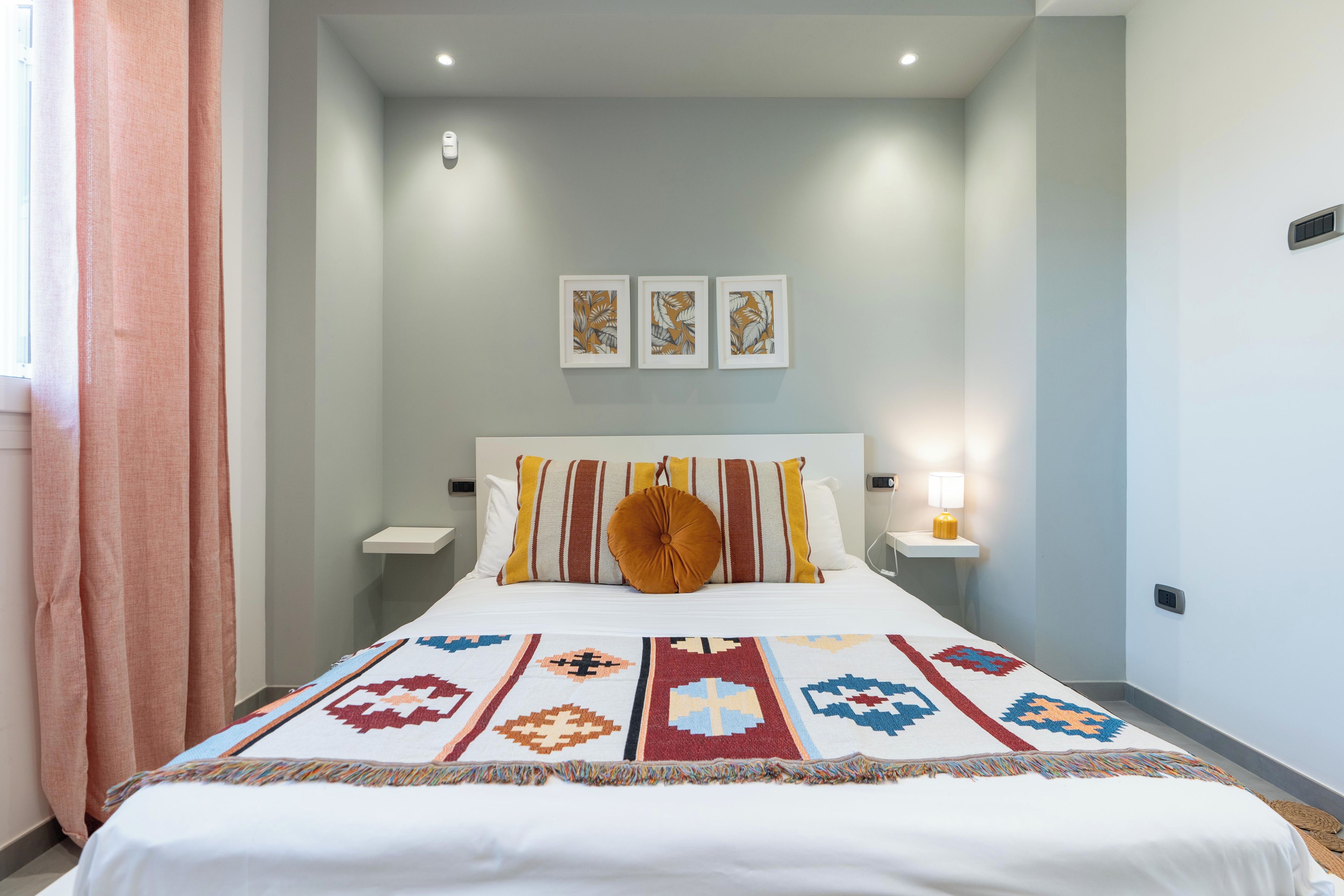 Un lit bien fait, recouvert d’une couverture à motifs, deux tables de chevet avec des lampes et trois œuvres d’art encadrées accrochées au mur.