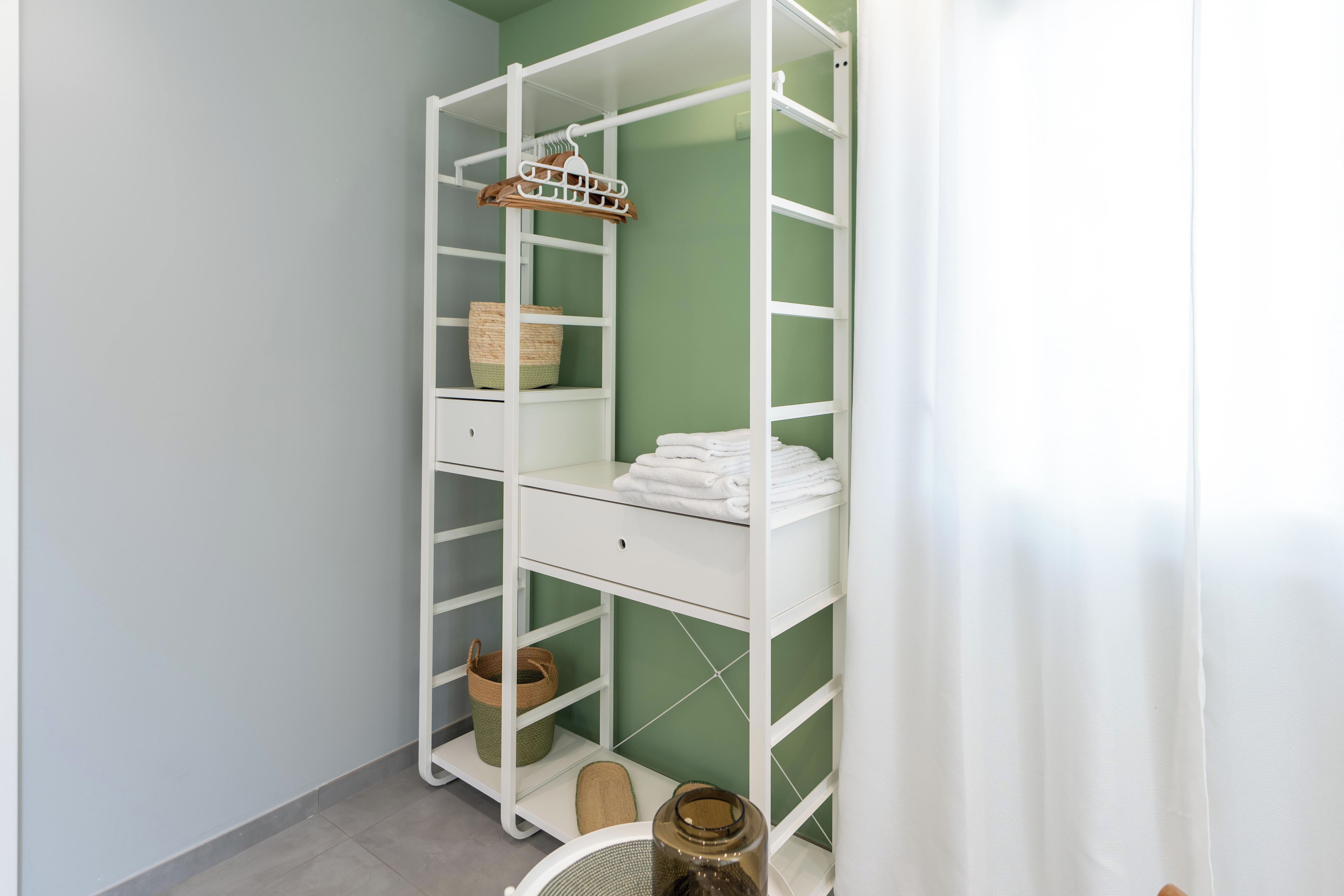 A white metal wardrobe with shelves and drawers, baskets, and a hanging rack against a green wall.