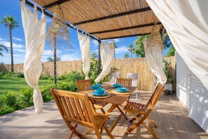 Un patio al aire libre con una mesa y sillas de madera, rodeado de cortinas blancas y un techo de bambú.