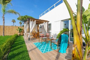 A patio with a wooden table and chairs, a blue patterned rug, and a pergola with white curtains. - Perla del Mare (Ispica)