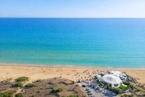 An aerial view of a beach with a large dome-shaped building, a parking area, and a sandy beach with umbrellas and people. - Perla del Mare (Ispica)