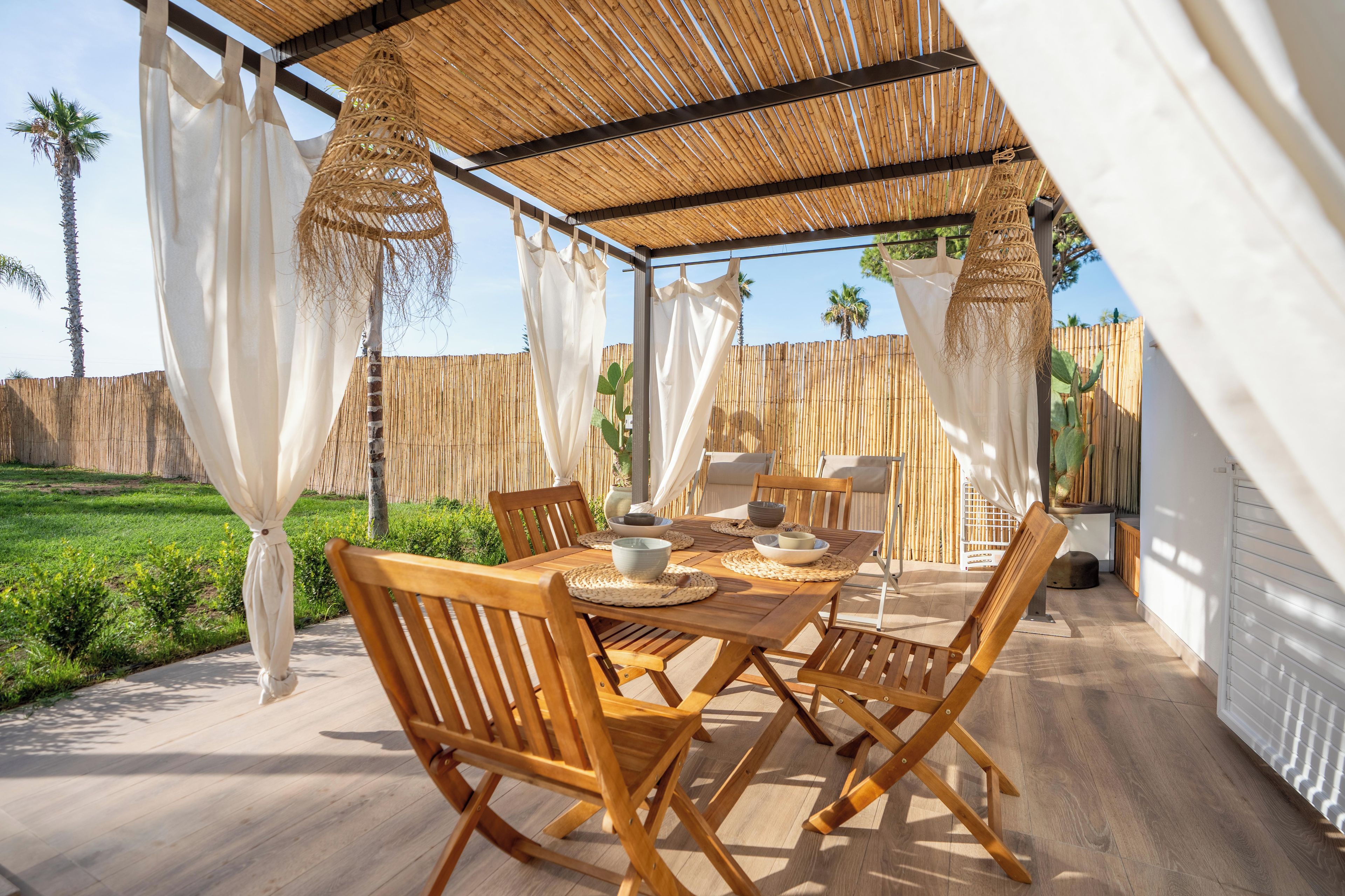 An outdoor dining area with a wooden table and chairs, a thatched roof, and a view of a garden.