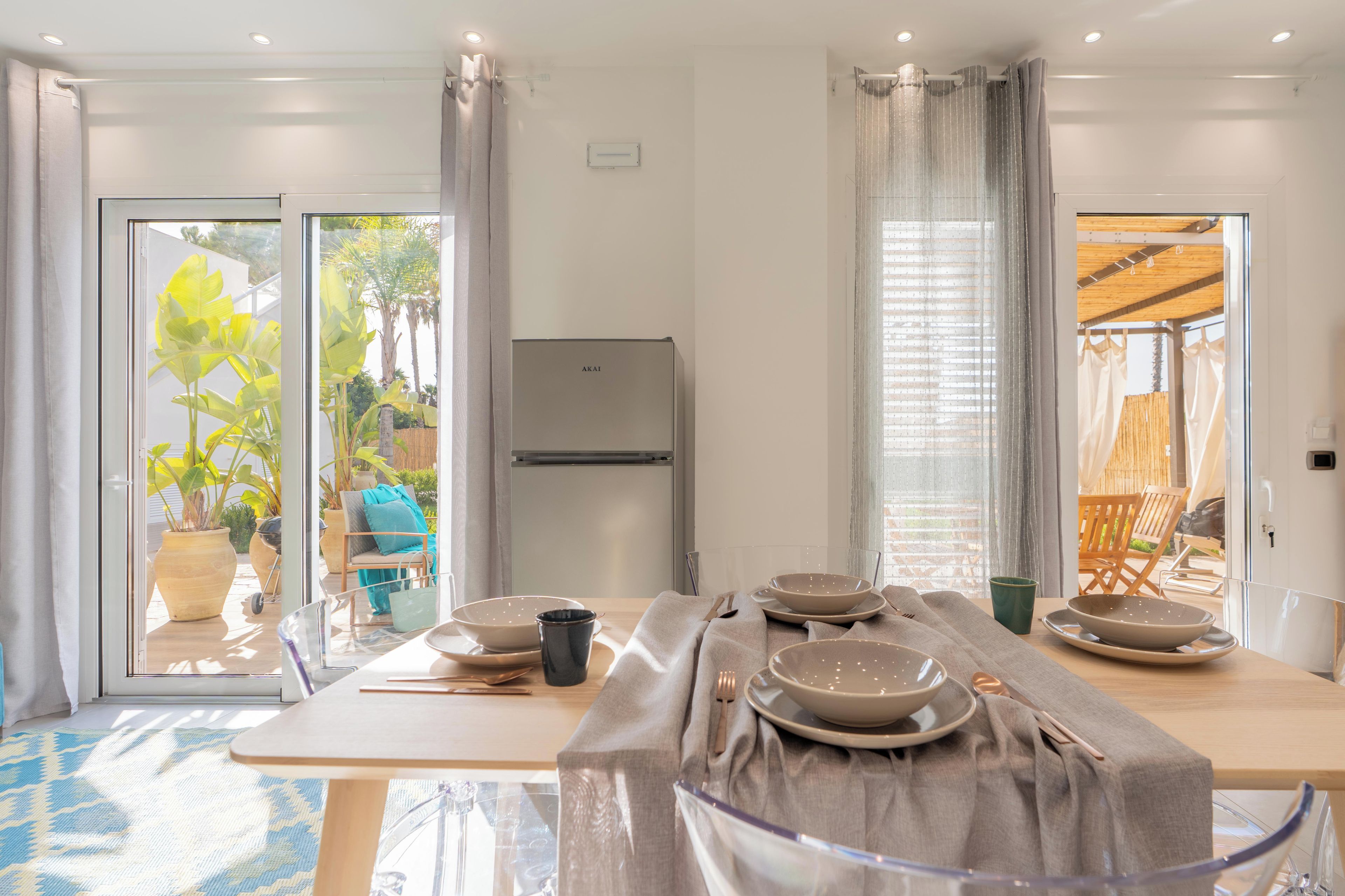 A dining area with a table set for four, a refrigerator, and a view of the garden through glass doors.