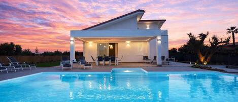 A modern house with a swimming pool, outdoor seating, and a sunset sky.