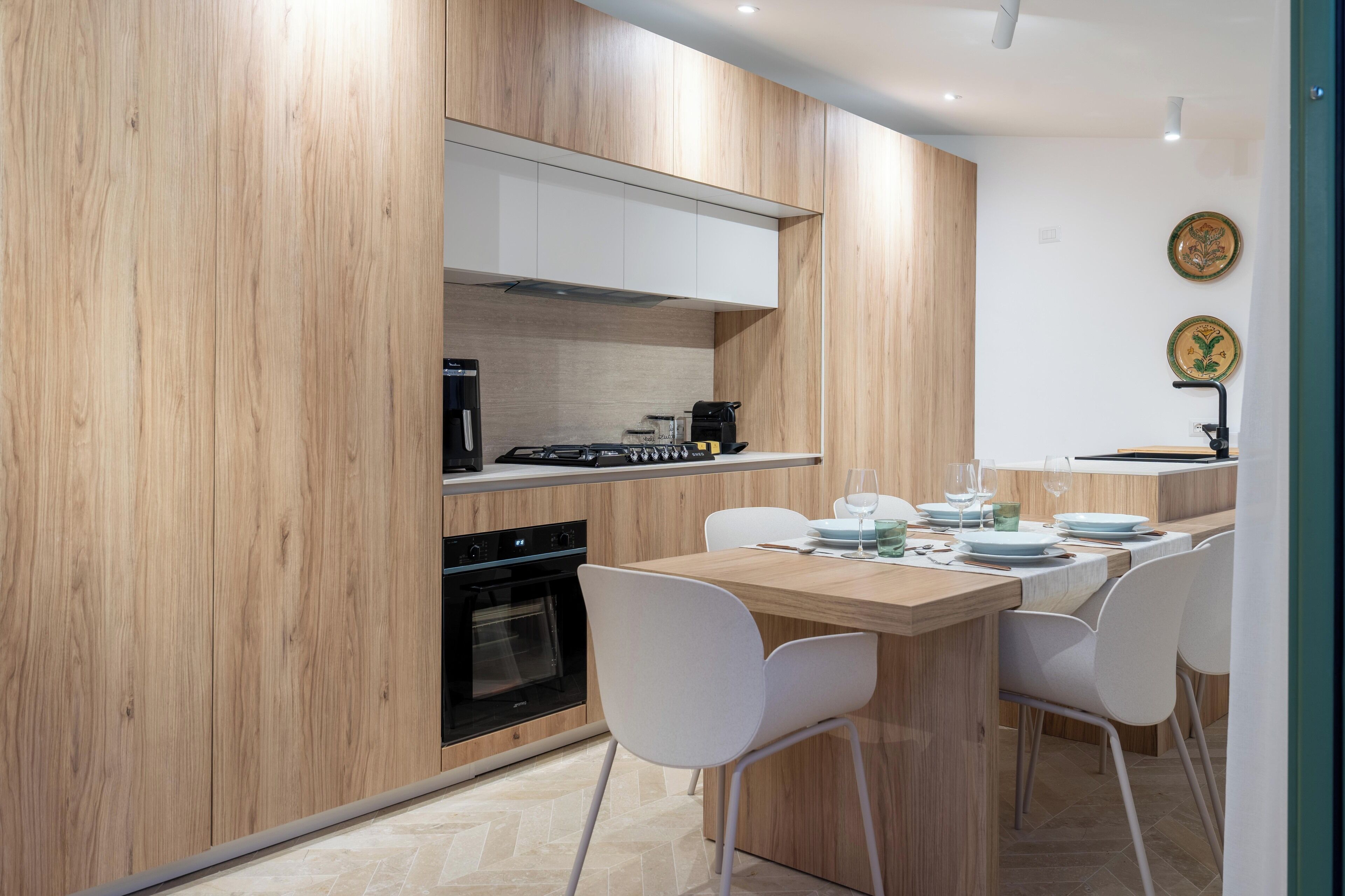 A modern kitchen with wooden cabinetry, a built-in oven, and a dining area with white chairs and a wooden table.