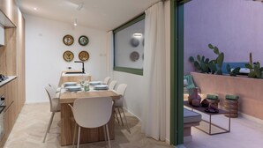 Un comedor moderno con una mesa de madera y sillas blancas, un área de cocina con armarios de madera, y vistas a un jardín de cactus a través de una puerta de cristal.