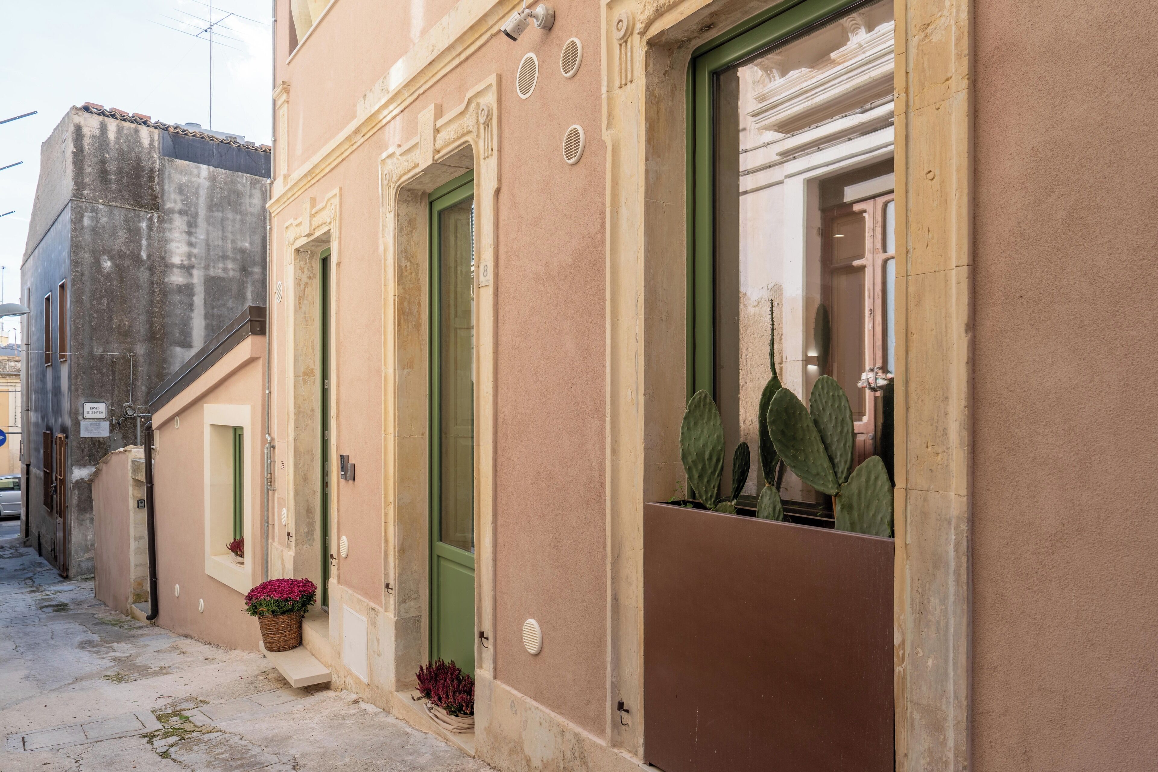 Una calle estrecha con un edificio que tiene puertas verdes y un pequeño balcón con plantas.
