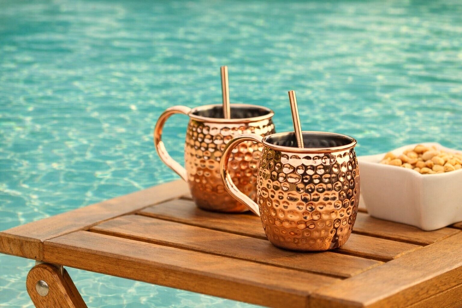 Two copper mugs with straws on a wooden table by a poolside.