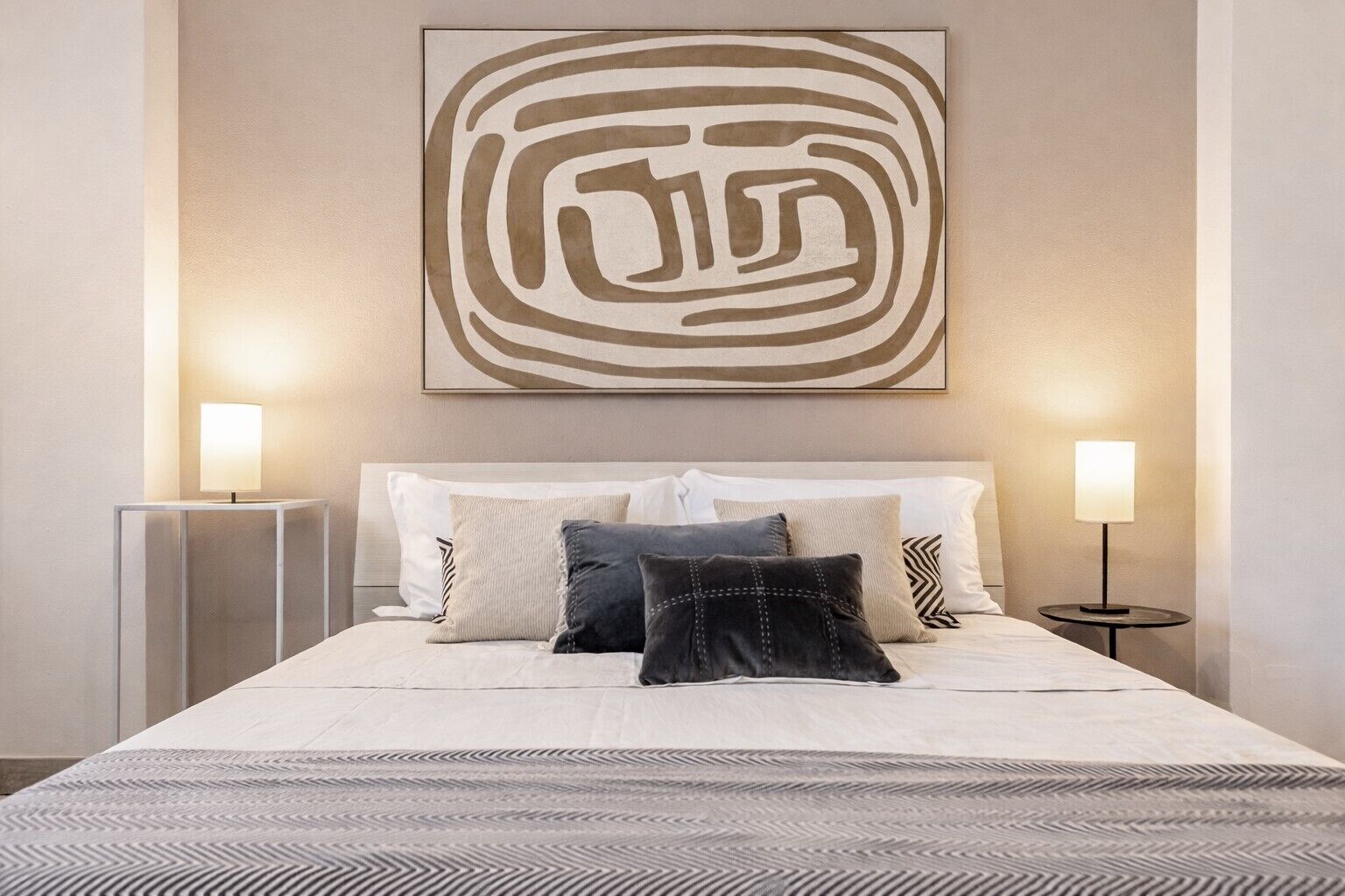 Een netjes opgemaakt bed met een beddengoed met patroon, twee bedlampjes en een groot abstract kunstwerk erboven.