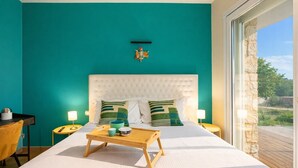 Une chambre avec des murs de couleur turquoise, une tête de lit blanche à motifs capitonnés, deux tables de chevet, une table de chevet en bois avec un plateau, et une vue sur l’extérieur grâce à une porte vitrée.