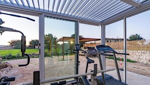 Un espace de remise en forme extérieur abrité, équipé de matériel d’exercice, notamment un tapis de course et des appareils de musculation, et offrant une vue sur un quartier résidentiel avec une voiture et une maison en arrière-plan.