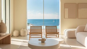 Ein modernes Wohnzimmer mit einem großen Schiebeglasfenster, das auf einen Balkon mit Meerblick führt. Zwei Korbstühle und ein runder Couchtisch sind vorhanden.