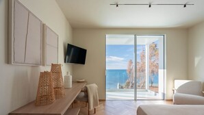 Ein modernes Schlafzimmer mit einem großen Bett, einem an der Wand montierten Fernseher, einem Holztischettchen und Meerblick durch eine Glastür.