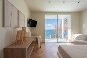 Ein modernes Schlafzimmer mit einem großen Bett, einem an der Wand montierten Fernseher, einem Holztischettchen und Meerblick durch eine Glastür.