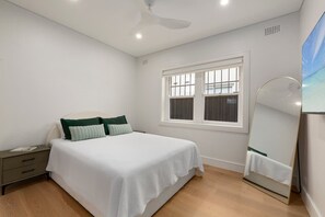 Quarto com cama, mesa de cabeceira, janela e ventoinha no teto.