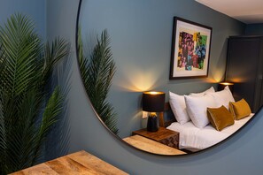 Un espejo curvo que refleja un dormitorio con una cama, almohadas, una mesita de noche, una lámpara y un cuadro en la pared.