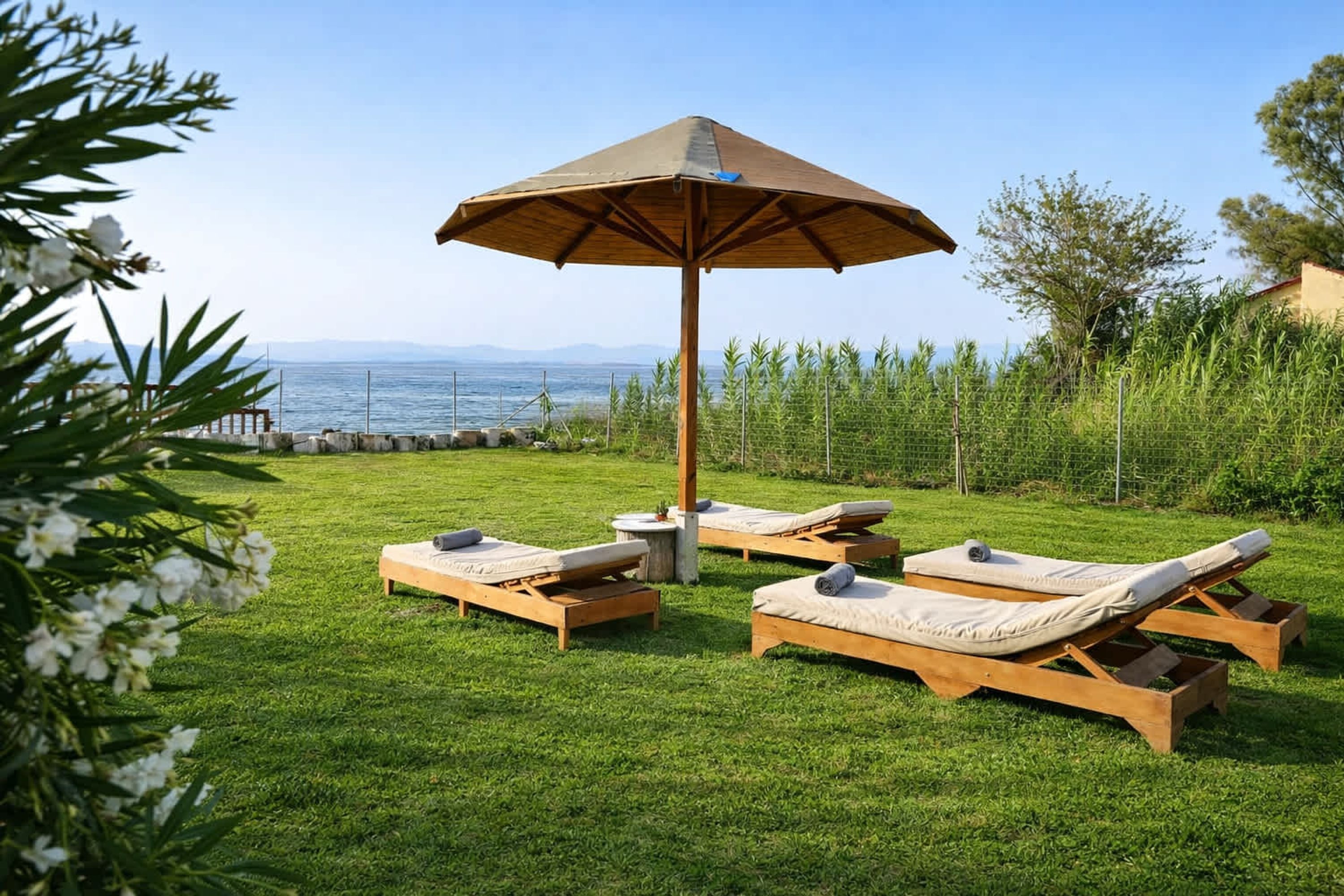 A grassy outdoor area with wooden lounge chairs and a large umbrella, overlooking a body of water.