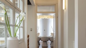 Un pasillo con suelo de madera, un jarrón blanco con flores y vistas a una habitación con un ventanal y un sofá.