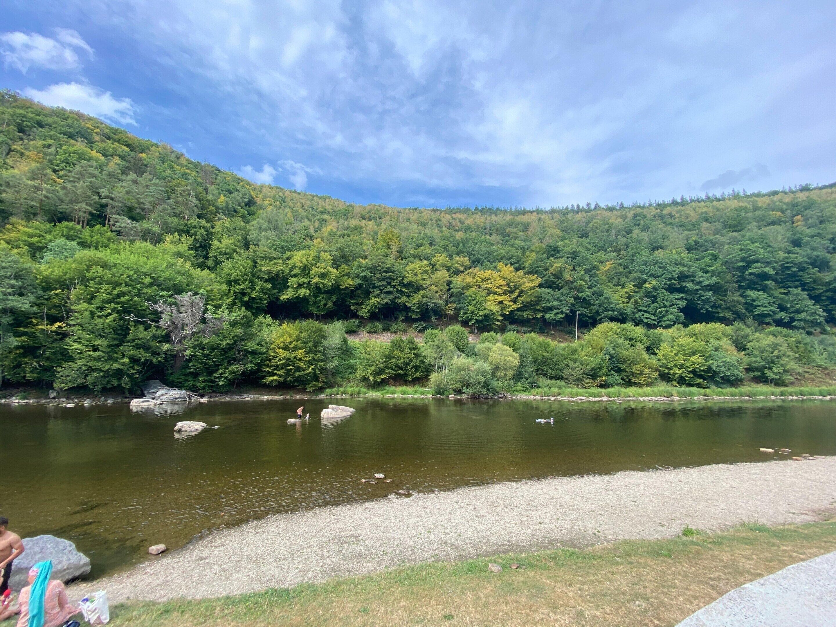 A river with a forested hillside, a gravel path, and a person by the water.
