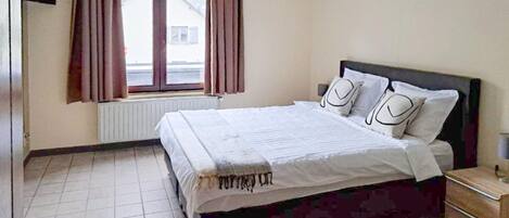 Quarto com cama, cortinas, radiador, janela e mesa de cabeceira.