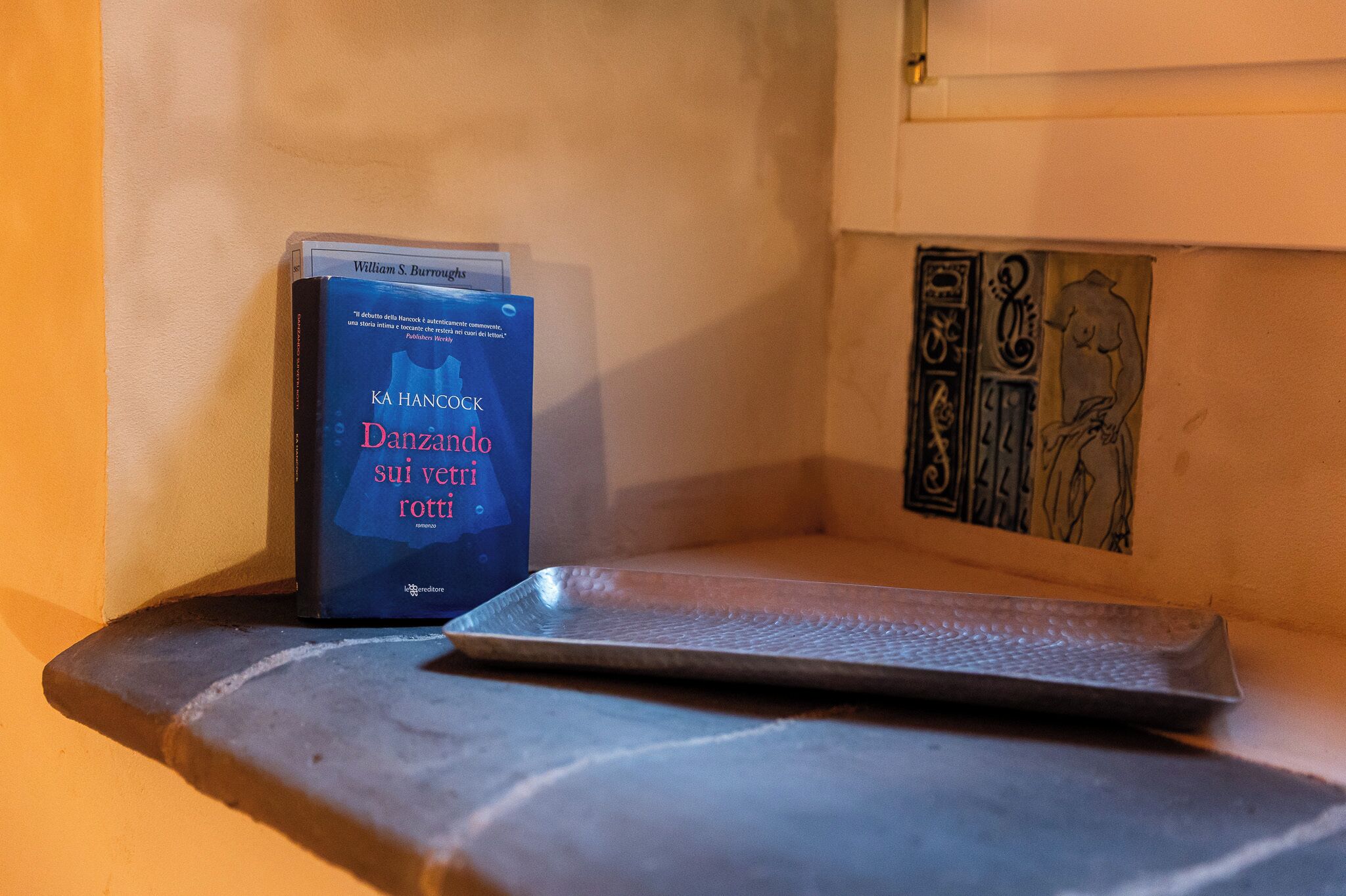 Ein Buch mit dem Titel 'Danzando sui venti rotti' auf einem Regal, eine Metalltablett und eine Wand mit einem Zierpaneel.