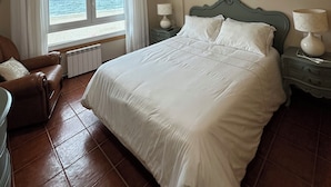 A hotel room with a large bed, two bedside tables with lamps, a view of the sea through a window, a brown leather chair, and a radiator under the window. - Unbeatable location, first line of the beach in the middle of Silgar promenade. (sanxenxo)