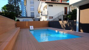Ein Schwimmbecken mit Holzterrasse, einem Nichtschwimmender-Schild und einem Gebäude mit Balkonen im Hintergrund.