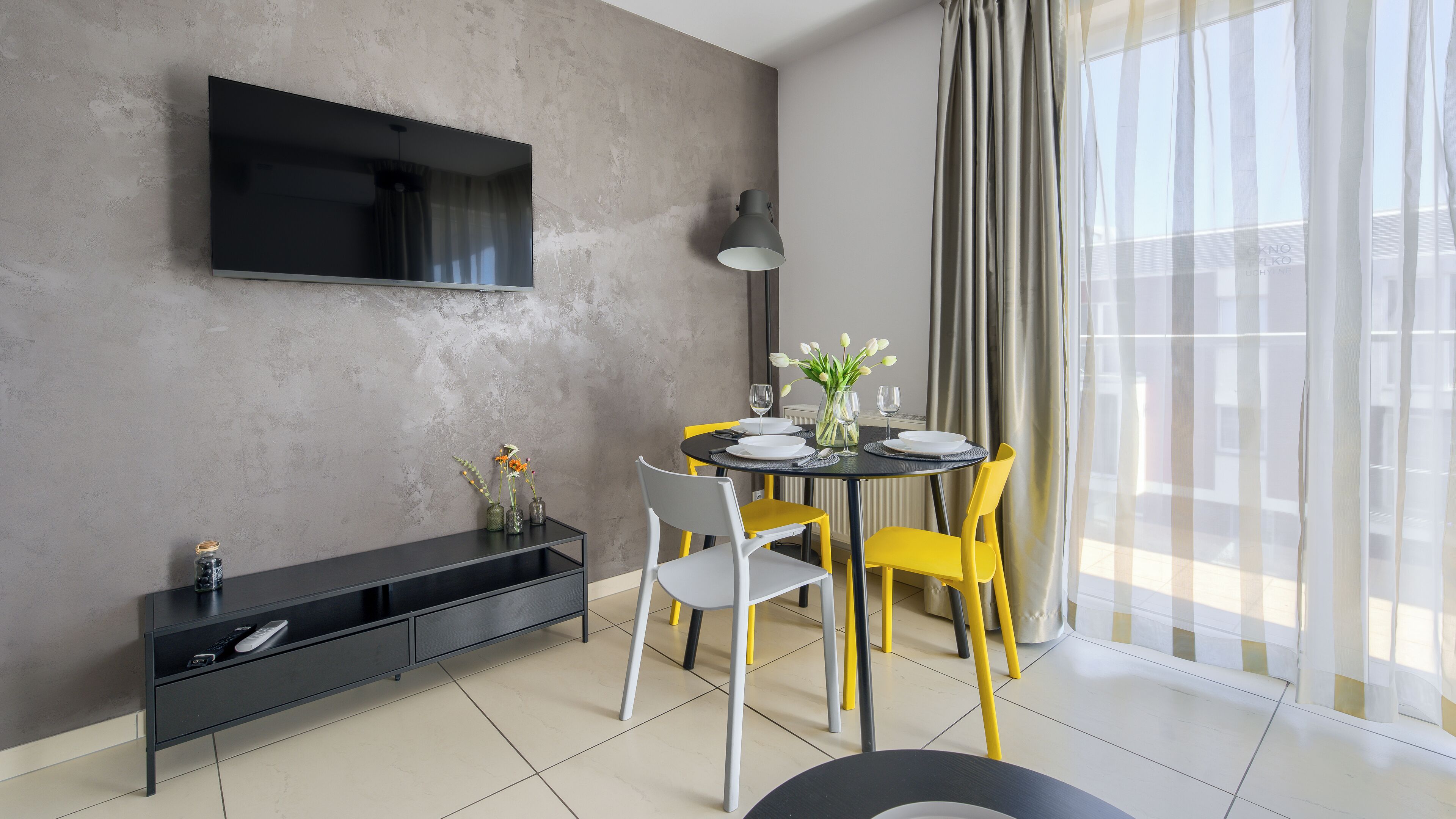 A modern dining area with a TV mounted on the wall, a dining table set with plates and glasses, yellow chairs, and a black console table with a vase of flowers.