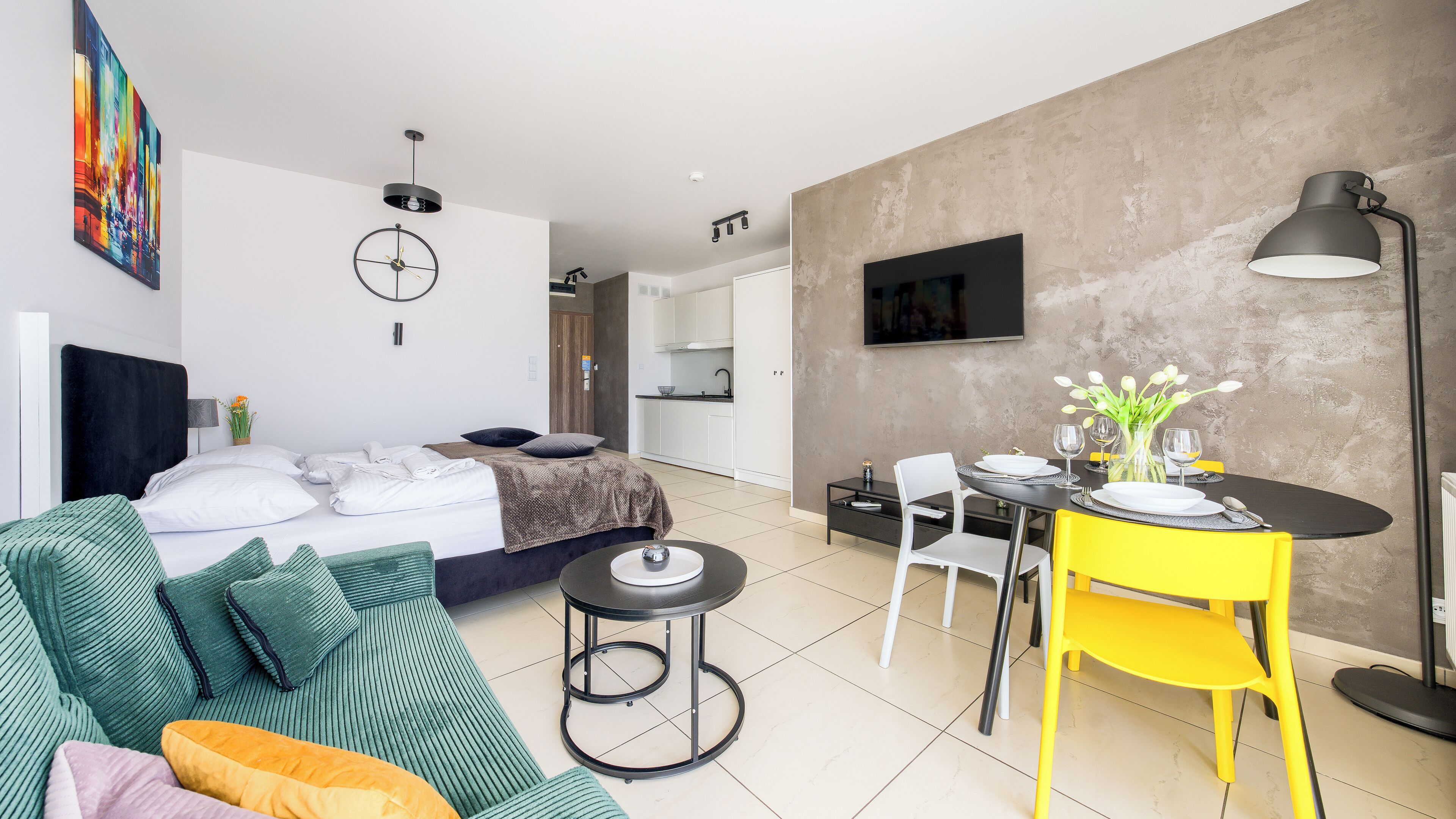 Une chambre d’hôtel moderne équipée d’un lit, d’un canapé, d’une table à manger avec des chaises, d’un téléviseur fixé au mur et d’un petit coin cuisine.