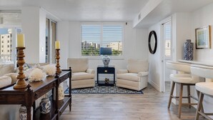 Ein Wohnzimmer mit einer Couch, Sesseln, einem Couchtisch und einem Teppich. Es gibt Fenster mit Jalousien, eine Lampe und eine Vase auf einem Beistelltisch.