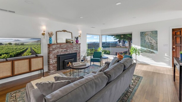 A modern living room with a fireplace, comfortable seating, and a large window offering a view of a vineyard. - Ocean Breeze-Charming, 3 bedroom 2 bath home in old town Bodega Bay (Bodega Bay)
