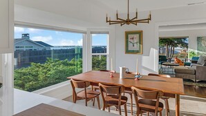 A dining area with a large table, chairs, and a chandelier. There are windows offering a view of greenery and buildings. - Ocean Breeze-Charming, 3 bedroom 2 bath home in old town Bodega Bay (Bodega Bay)