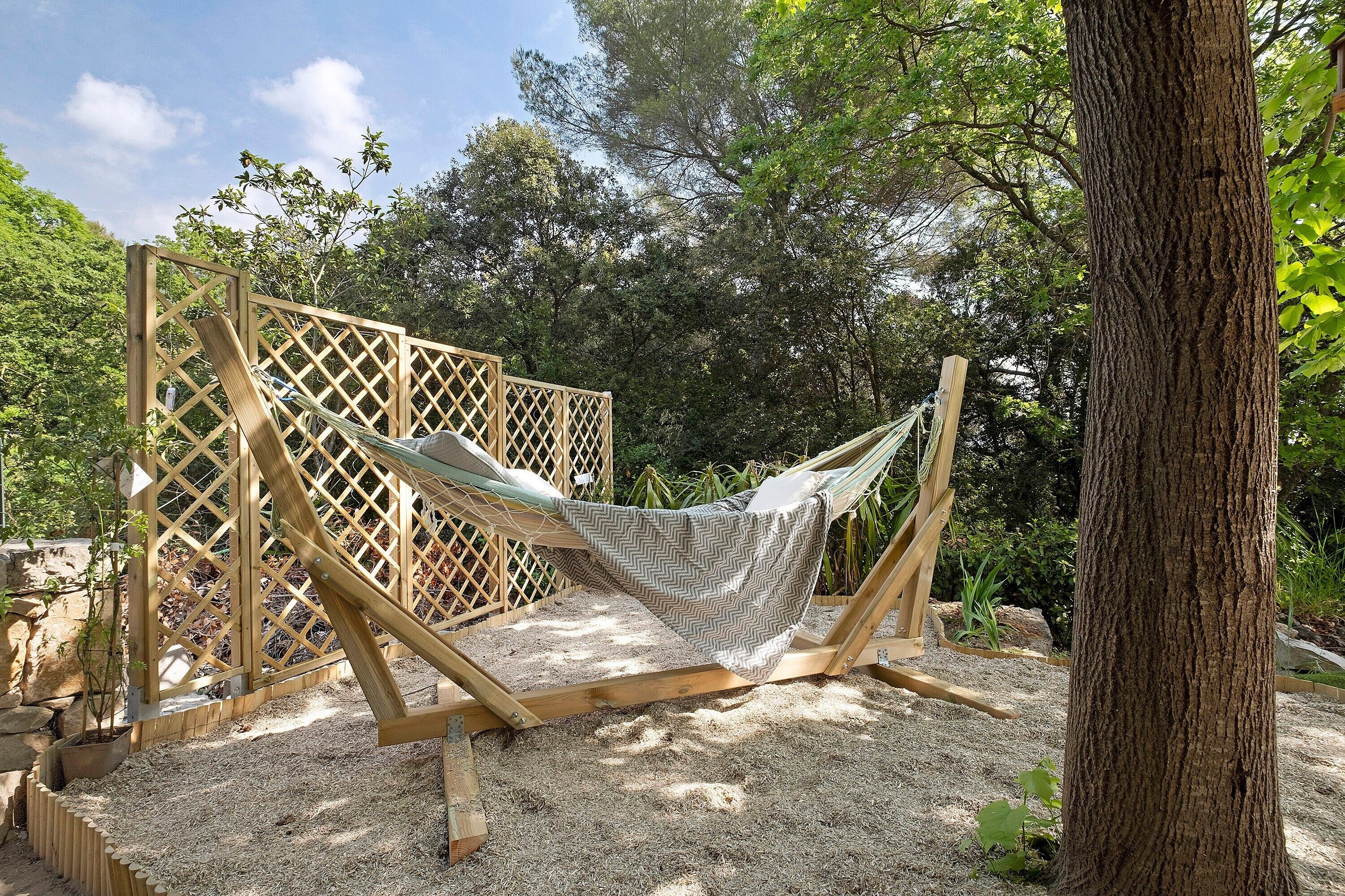 Una hamaca colocada entre paneles de madera en un jardín con árboles y el tronco de un árbol a la vista.