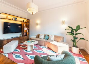Ein modernes Wohnzimmer mit Ecksofa, Couchtisch, Fernseher, Bücherregal und einer großen Pflanze.