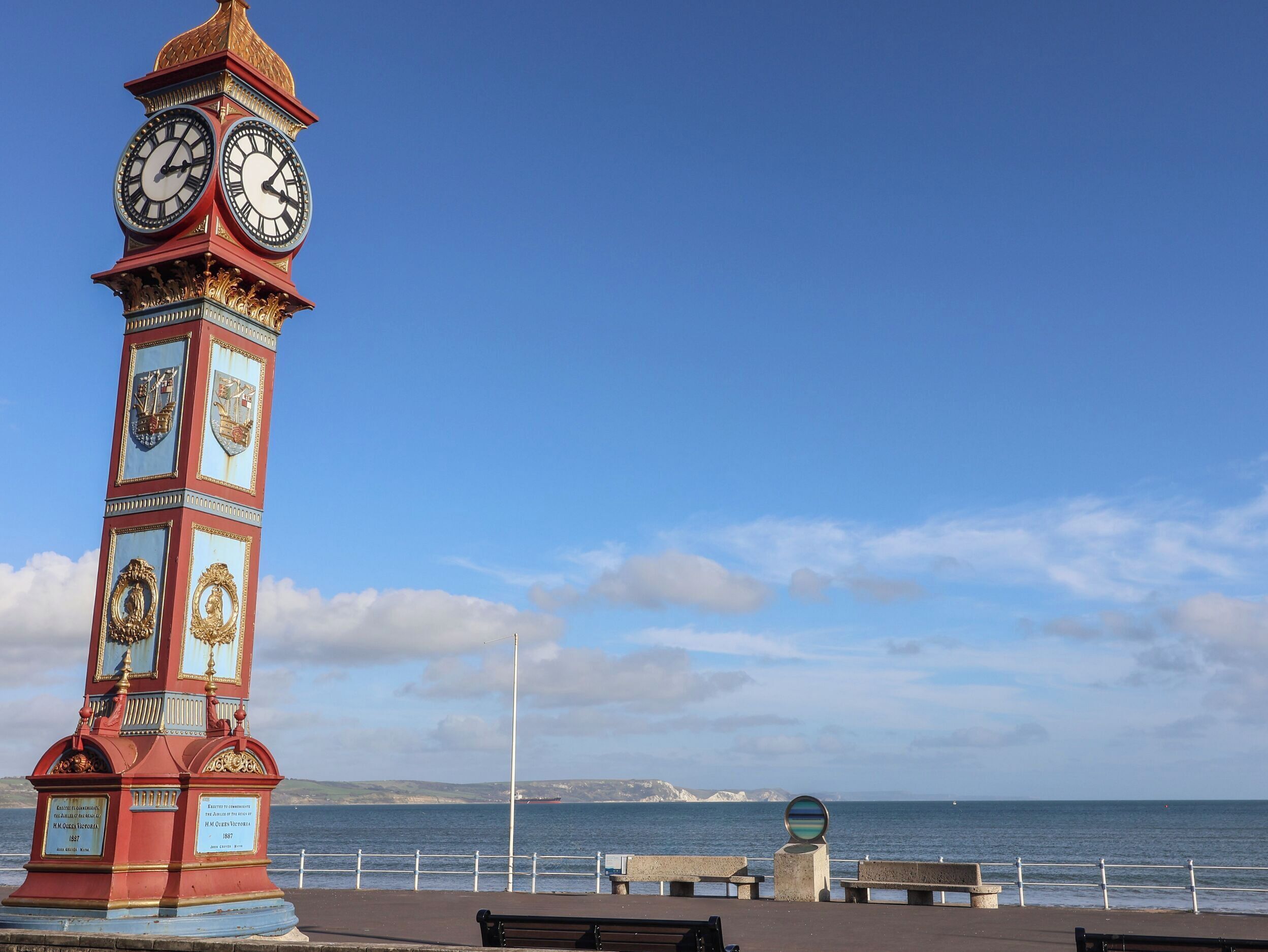 A clock tower with multiple faces, located near a waterfront with benches and a railing.
