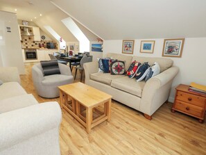 A spacious living area with a sofa, armchairs, a wooden coffee table, and a dining table with chairs. There is a kitchen in the background, and the room has a sloped ceiling with skylights. - Harbour Reach 7 (Weymouth)