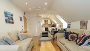 A cozy living room with a sofa, armchair, and coffee table. A kitchen is visible in the background with a dining table and chairs. - Harbour Reach 7 (Weymouth)
